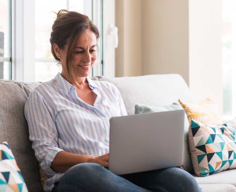 Middle aged woman using laptop in the sofa with a happy face standing and smiling with a confident smile showing teeth