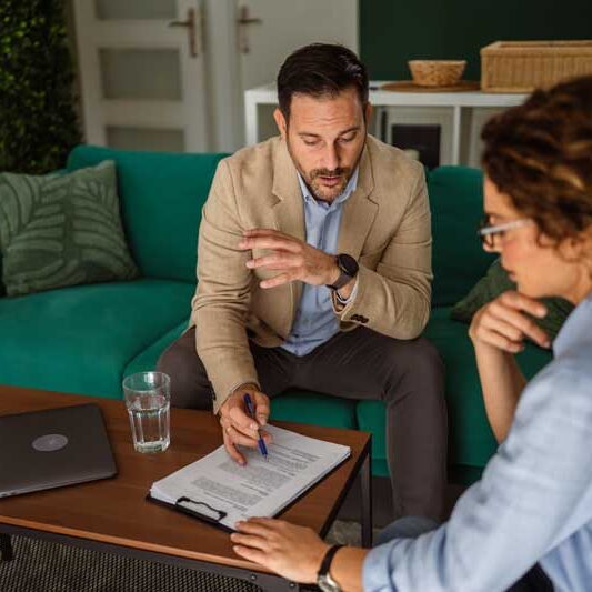 Financial adviser reviews agreement details with a woman at a modern casual office desk, offering guidance and support before signing paperwork to plan investments and partnerships